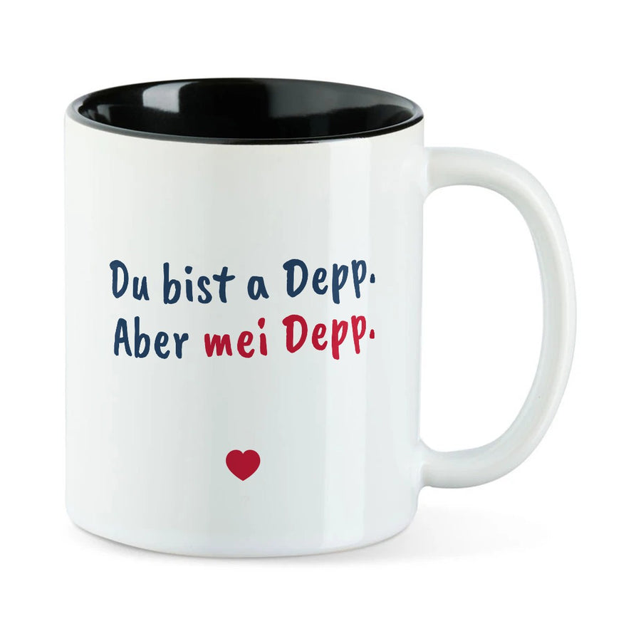 Weiße Tasse mit schwarzer Innenseite und dem bairischen Spruch „Du bist a Depp. Aber mei Depp.“ in Pinselschrift.
