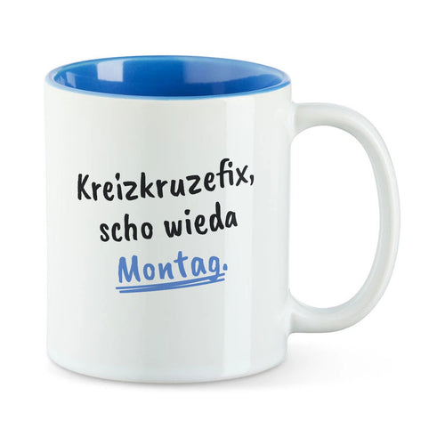 Weiße Tasse mit blauer Innenseite und dem bairischen Spruch „Kreizkruzefix, scho wieda Montag.“ in Pinselschrift.