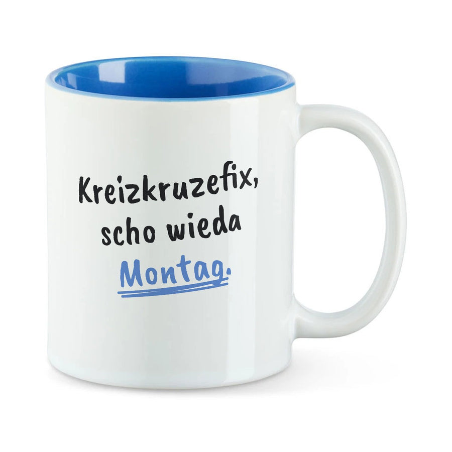 Weiße Tasse mit blauer Innenseite und dem bairischen Spruch „Kreizkruzefix, scho wieda Montag.“ in Pinselschrift.