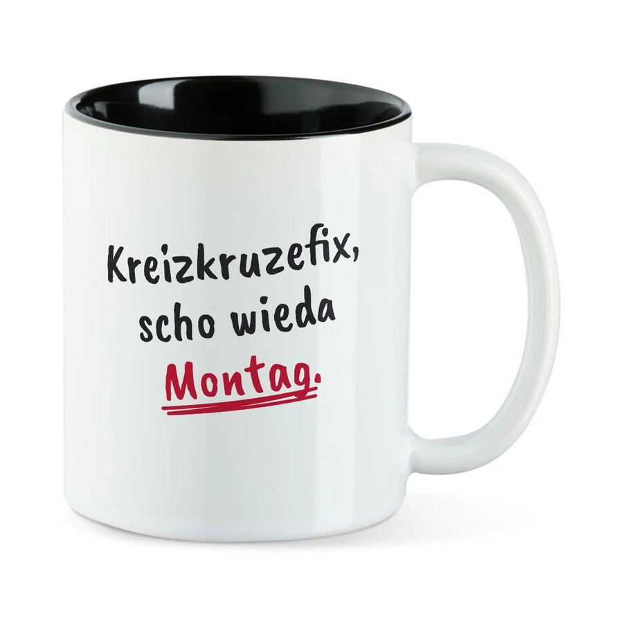 Weiße Tasse mit schwarzer Innenseite und dem bairischen Spruch „Kreizkruzefix, scho wieda Montag.“ in Pinselschrift.