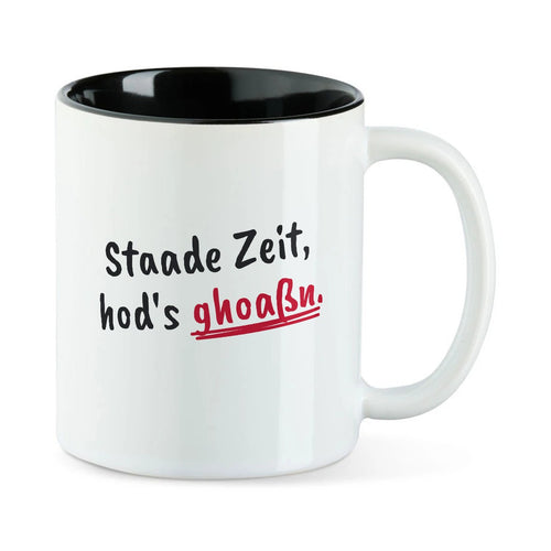 Weiße Tasse mit schwarzer Innenseite und dem bairischen Spruch „Staade Zeit, hod's ghoaßn.“ in Pinselschrift.
