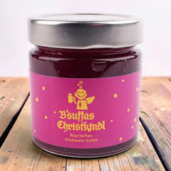 Glas B'suffas Christkindl, Glühwein-Gelee, pinkes Etikett mit lachendem Engel mit Krug und Sternen, auf einem Holztisch.