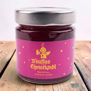 Glas B'suffas Christkindl, Glühwein-Gelee, pinkes Etikett mit lachendem Engel mit Krug und Sternen, auf einem Holztisch.