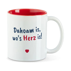 Weiße Tasse mit roter Innenseite und dem bairischen Spruch „Dahoam is, wo's Herz is!“ in Pinselschrift.