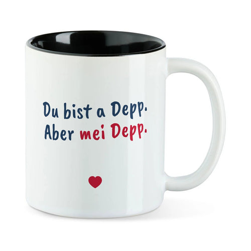 Weiße Tasse mit schwarzer Innenseite und dem bairischen Spruch „Du bist a Depp. Aber mei Depp.“ in Pinselschrift.