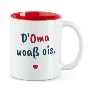 Weiße Tasse mit roter Innenseite und dem bairischen Spruch „D'Oma woaß ois.“ in Pinselschrift.