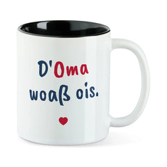 Weiße Tasse mit schwarzer Innenseite und dem bairischen Spruch „D'Oma woaß ois.“ in Pinselschrift.