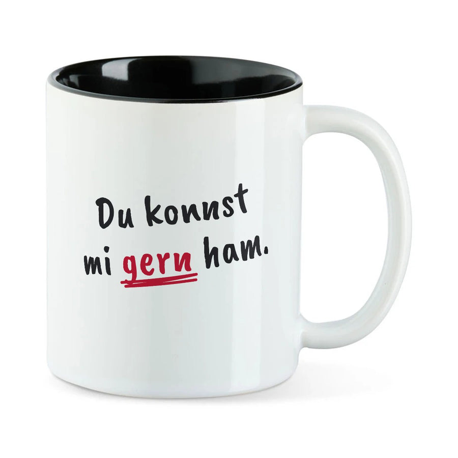 Weiße Tasse mit schwarzer Innenseite und dem bairischen Spruch „Du konnst mi gern ham.“ in Pinselschrift.