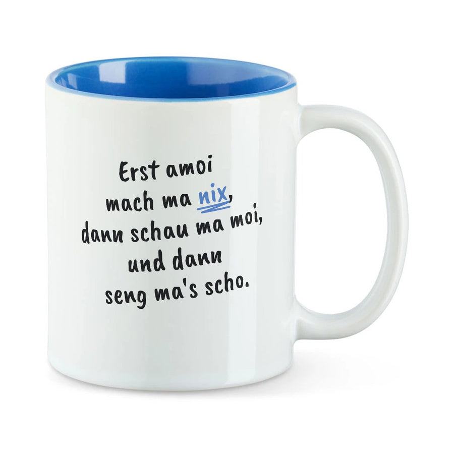 Weiße Tasse mit blauer Innenseite und dem bairischen Spruch „Erst amoi mach ma nix, dann schau ma moi, und dann seng ma's scho.“ in Pinselschrift.