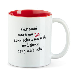 Weiße Tasse mit roter Innenseite und dem bairischen Spruch „Erst amoi mach ma nix, dann schau ma moi, und dann seng ma's scho.“ in Pinselschrift.