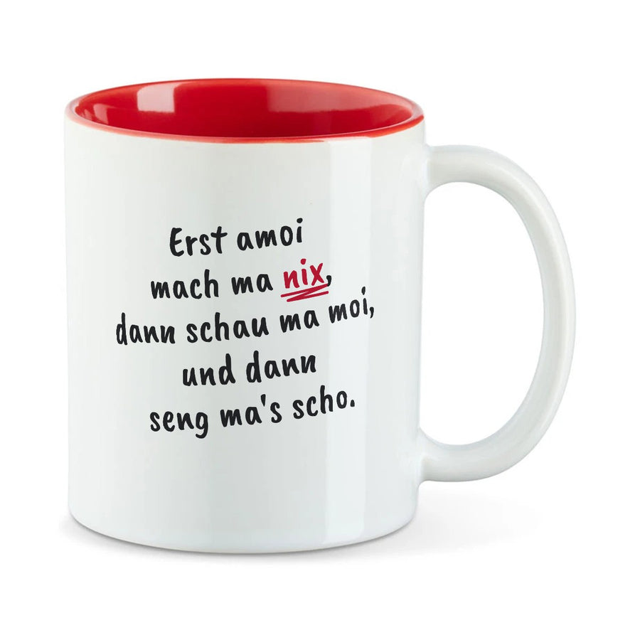Weiße Tasse mit roter Innenseite und dem bairischen Spruch „Erst amoi mach ma nix, dann schau ma moi, und dann seng ma's scho.“ in Pinselschrift.