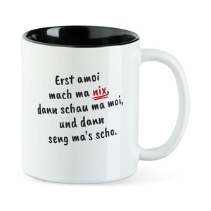 Weiße Tasse mit schwarzer Innenseite und dem bairischen Spruch „Erst amoi mach ma nix, dann schau ma moi, und dann seng ma's scho.“ in Pinselschrift.