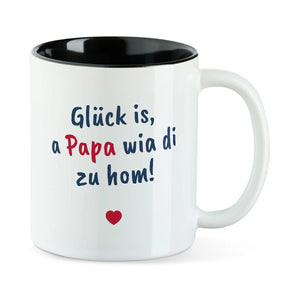 Weiße Tasse mit schwarzer Innenseite und dem bairischen Spruch „Glück is, a Papa wia di zu hom!“ in Pinselschrift.