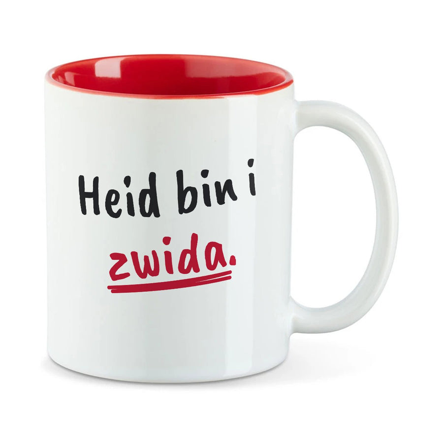 Weiße Tasse mit roter Innenseite und dem bairischen Spruch „Heid bin i zwida.“ in Pinselschrift.