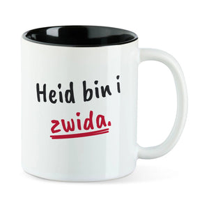 Weiße Tasse mit schwarzer Innenseite und dem bairischen Spruch „Heid bin i zwida.“ in Pinselschrift.