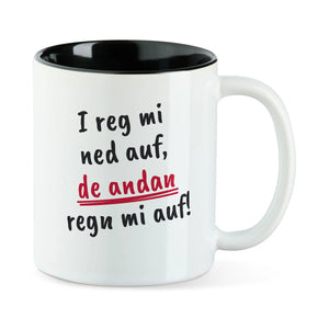 Weiße Tasse mit schwarzer Innenseite und dem bairischen Spruch „I reg mi ned auf, de andan regn mi auf!“ in Pinselschrift.