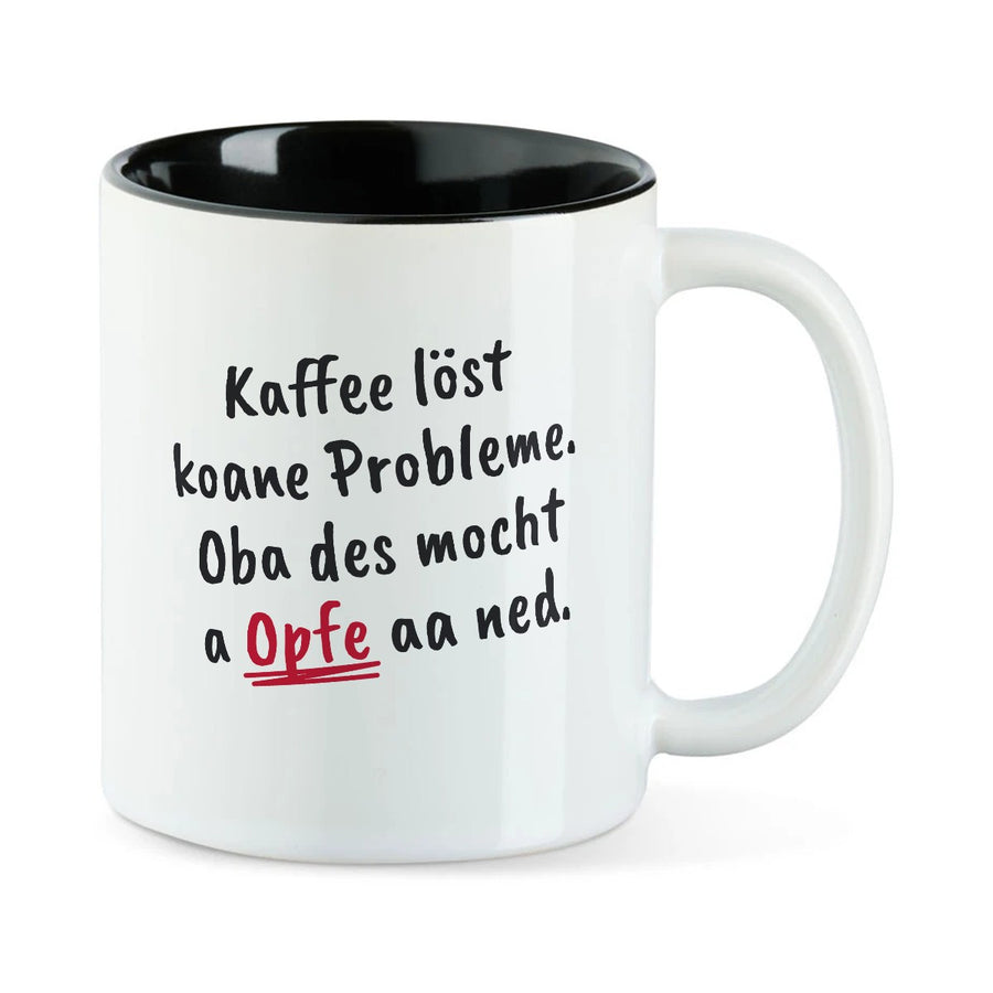 Weiße Tasse mit schwarzer Innenseite und dem bairischen Spruch „Kaffee löst koane Probleme. Oba des mocht a Opfe aa ned.“ in Pinselschrift.