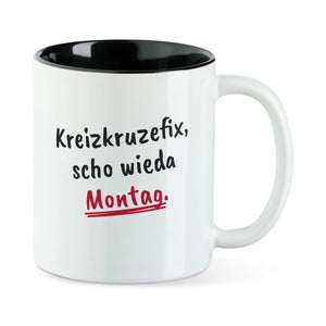 Weiße Tasse mit schwarzer Innenseite und dem bairischen Spruch „Kreizkruzefix, scho wieda Montag.“ in Pinselschrift.