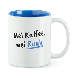 Weiße Tasse mit blauer Innenseite und dem bairischen Spruch „Mei Kaffee, mei Ruah.“ in Pinselschrift.