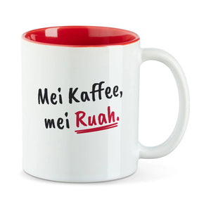 Weiße Tasse mit roter Innenseite und dem bairischen Spruch „Mei Kaffee, mei Ruah.“ in Pinselschrift.