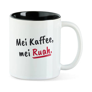 Weiße Tasse mit schwarzer Innenseite und dem bairischen Spruch „Mei Kaffee, mei Ruah.“ in Pinselschrift.