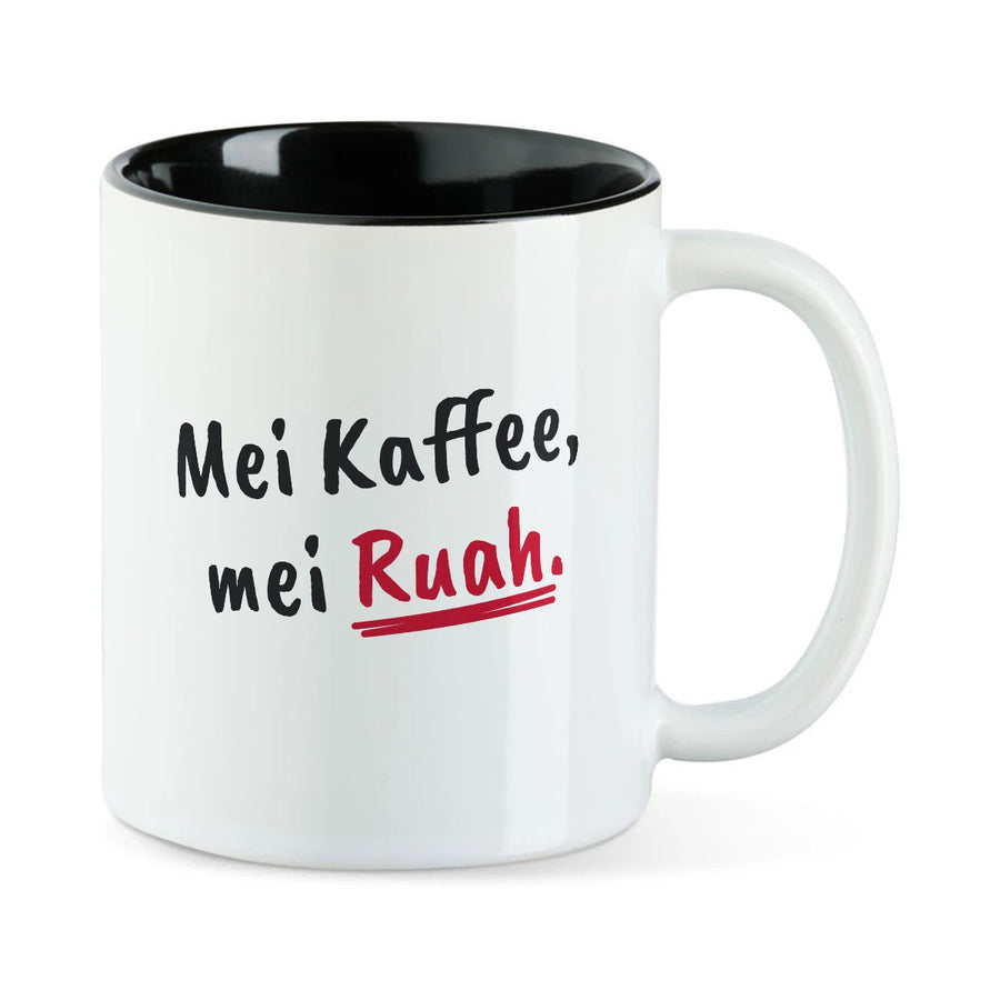 Weiße Tasse mit schwarzer Innenseite und dem bairischen Spruch „Mei Kaffee, mei Ruah.“ in Pinselschrift.