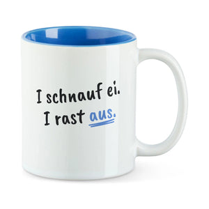 Weiße Tasse mit blauer Innenseite und dem bairischen Spruch „I schnauf ei. I rast aus.“ in Pinselschrift.