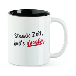 Weiße Tasse mit schwarzer Innenseite und dem bairischen Spruch „Staade Zeit, hod's ghoaßn.“ in Pinselschrift.