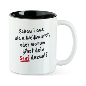 Weiße Tasse mit schwarzer Innenseite und dem bairischen Spruch „Schau i aus wia a Weißwurst, oder warum gibst dein Senf dazua!?“ in Pinselschrift.